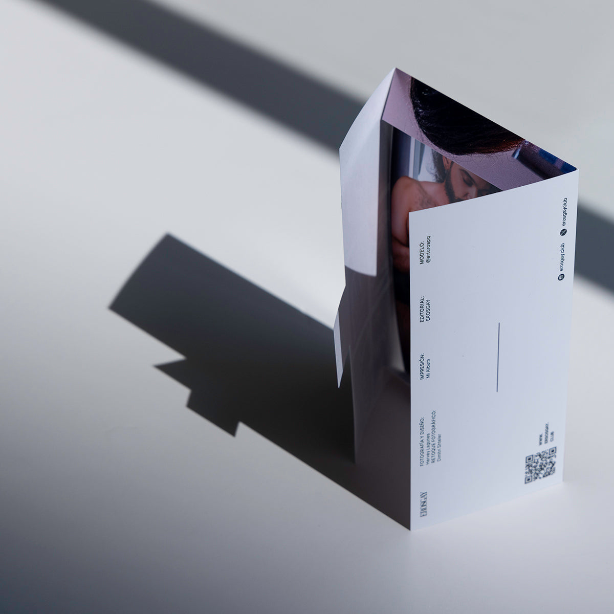 Vista lateral/trasera del empaque abierto del Sixpack de Arturo de ErosGay; se aprecia el panel blanco con créditos editoriales, redes sociales y un código QR, fotografiado sobre fondo blanco con una sombra diagonal marcada.
