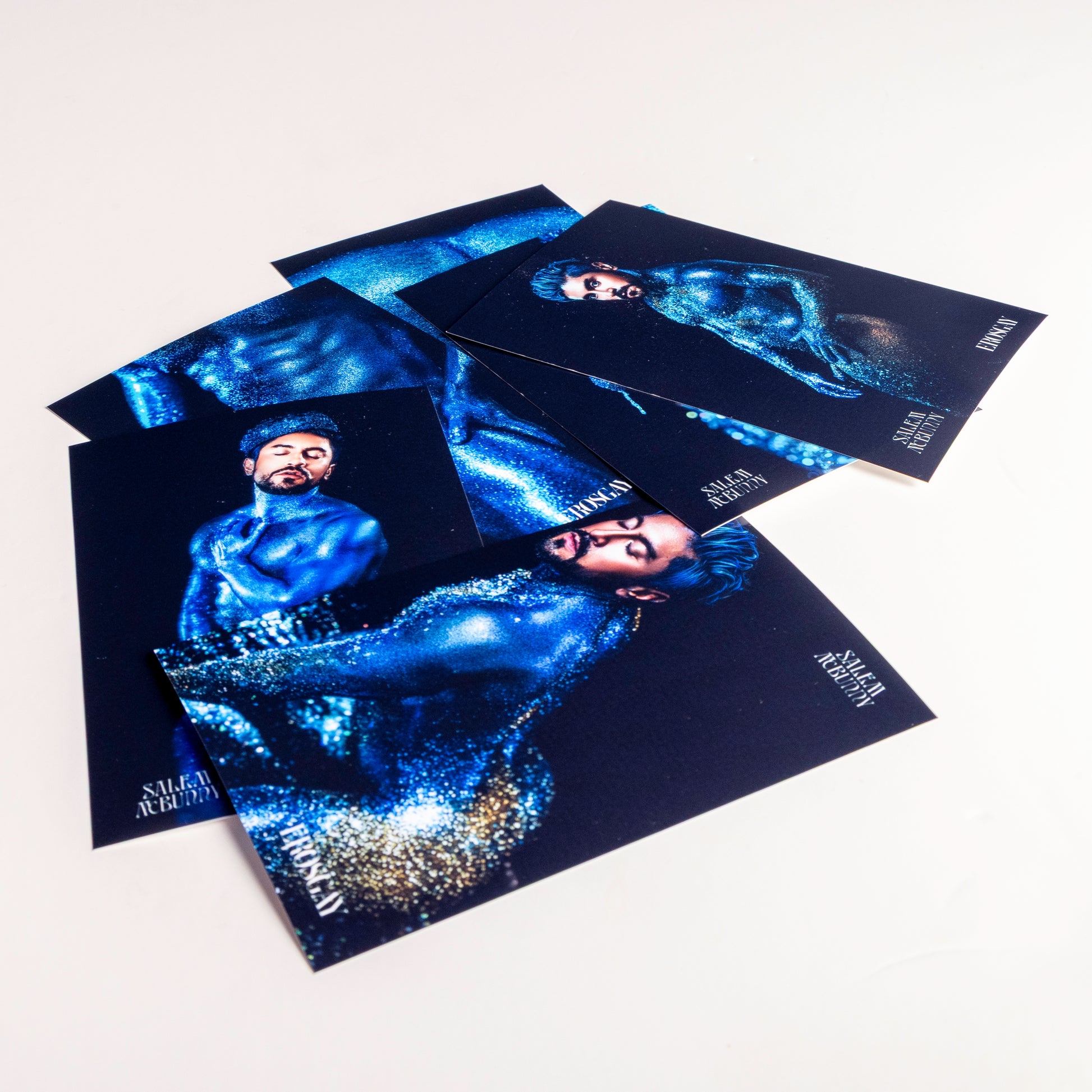 Seis impresiones fotográficas del Sixpack Oráculo (ErosGay) extendidas y traslapadas sobre un fondo blanco. Las imágenes muestran al modelo FelixFox cubierto de pigmento azul brillante en distintas poses