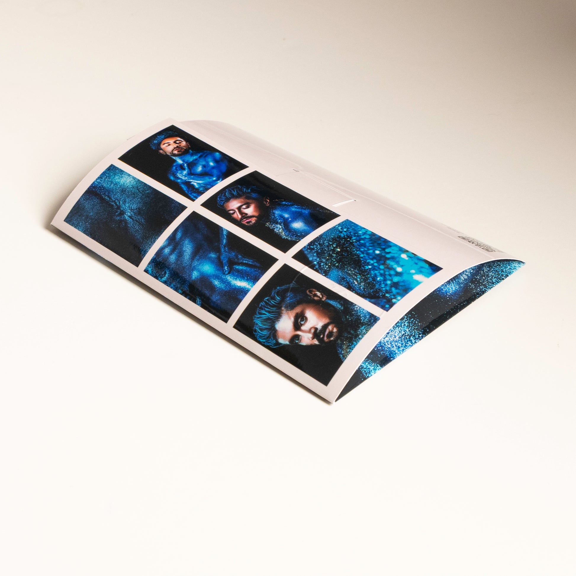 Vista trasera del sobre del producto Sixpack de ErosGay (colección Oráculo, modelo FelixFox), mostrando el reverso con la cuadrícula de 6 miniaturas/ventanas de la serie en tonos azules, sobre una superficie clara