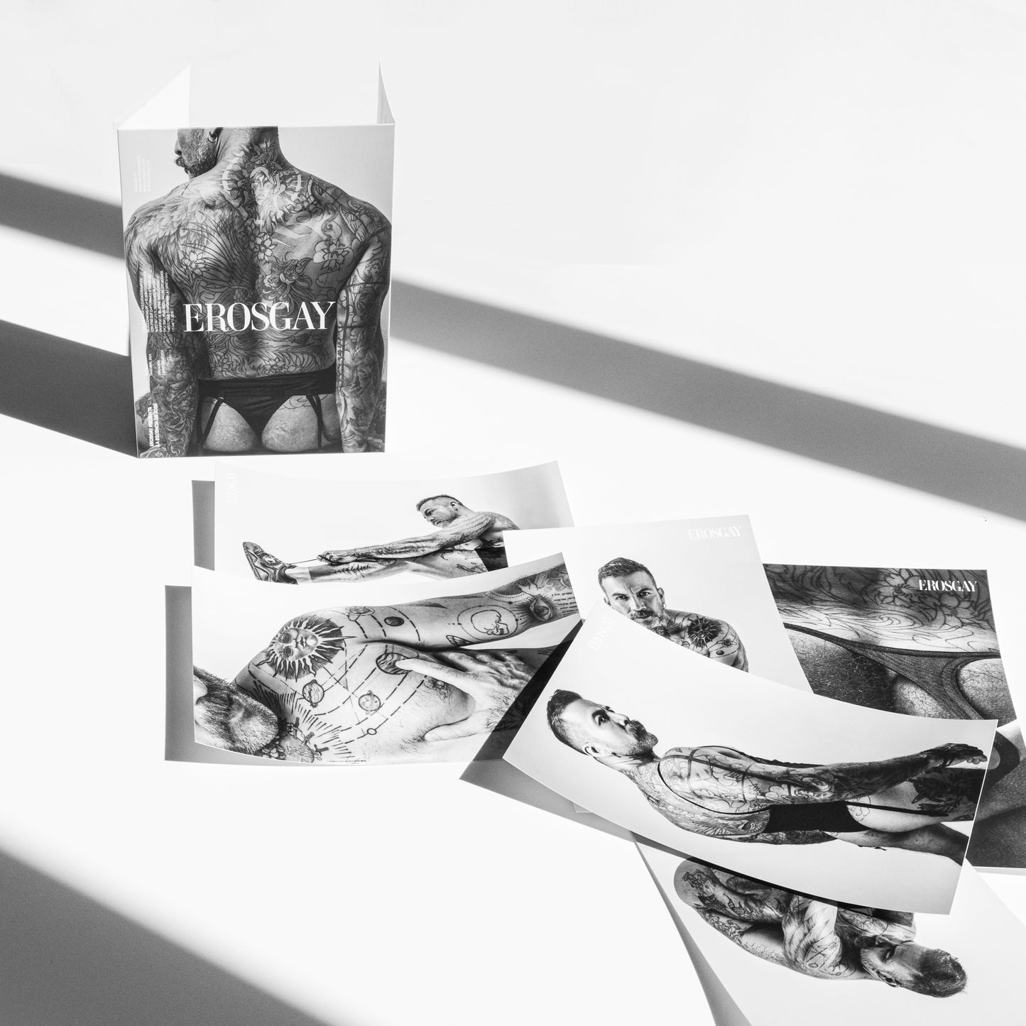 Sixpack de José de la colección “Tatuajes que hablan” de ErosGay con el contenido afuera: el sobre/estuche en blanco y negro a un lado y seis impresiones extendidas sobre fondo blanco, con sombras diagonales marcadas.