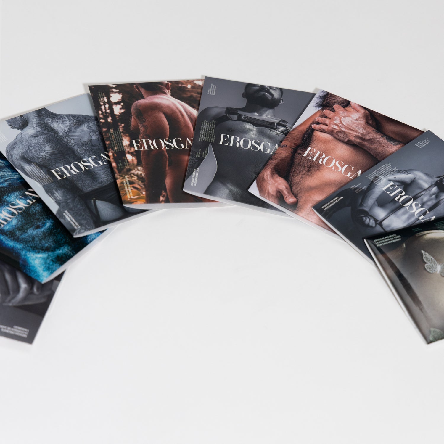Ocho sobres de sixpack ErosGay acomodados en arco sobre fondo blanco, mostrando diferentes portadas editoriales y dejando amplio espacio negativo al centro
