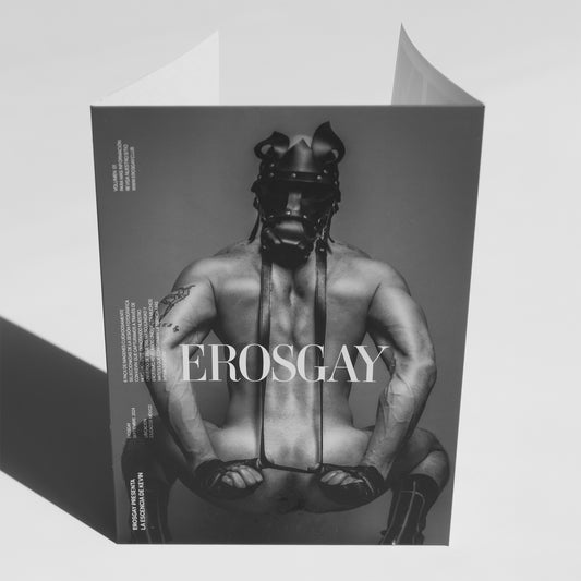 Sobre del sixpack de Kevin (ErosGay) en blanco y negro, fotografiado de frente sobre fondo gris claro. En la portada se ve al modelo de espaldas, usando un arnés y una máscara de cuero, con postura firme y estética editorial. El logotipo ‘EROSGAY’ aparece grande al centro. Se aprecian pequeños textos editoriales en el margen izquierdo y las solapas superiores del sobre ligeramente abiertas, proyectando sombras