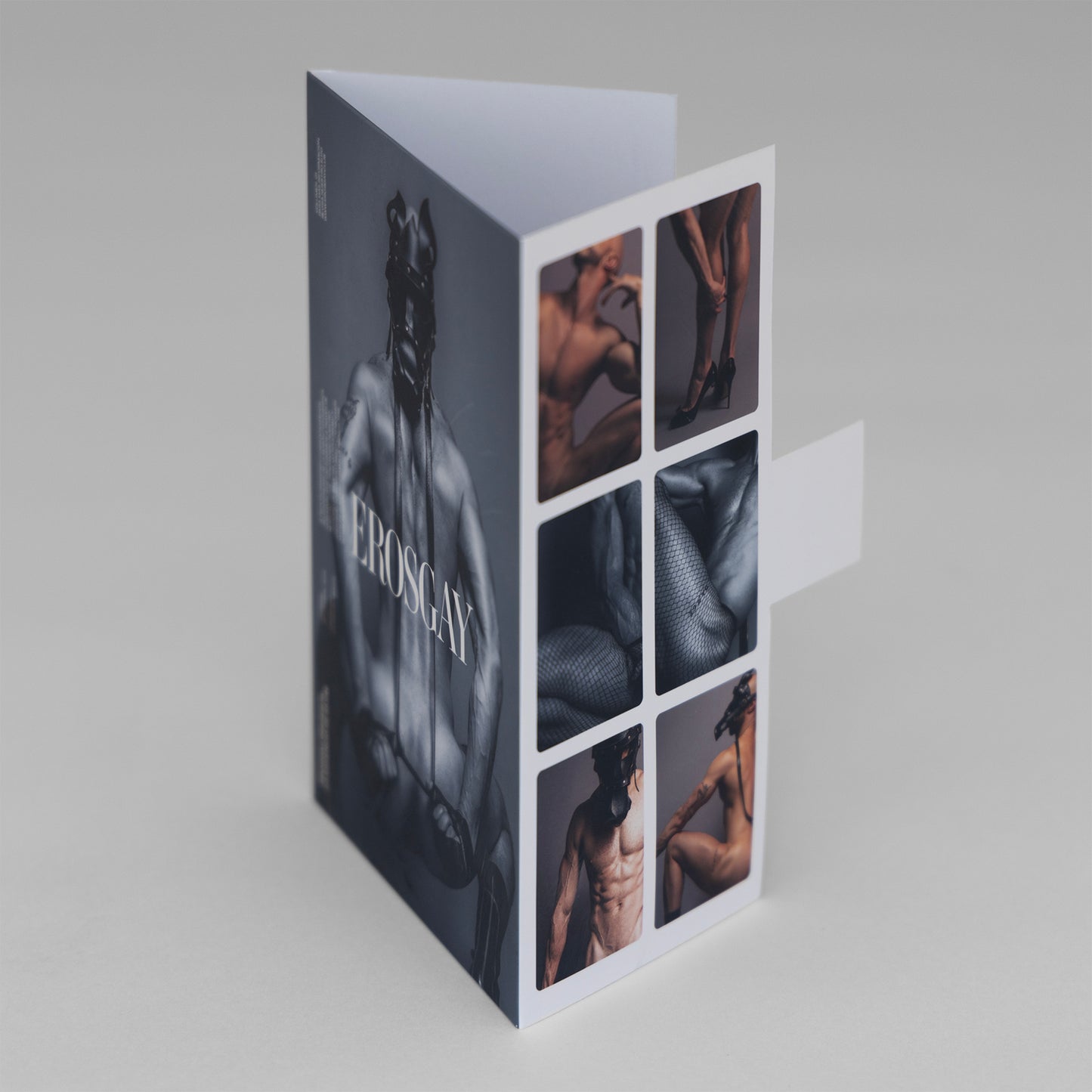 Vista lateral del sobre del sixpack de Kevin (ErosGay) sobre fondo gris. Se aprecia el lomo y parte de la portada en blanco y negro con el logo ‘EROSGAY’ en vertical, mientras que el panel trasero muestra una retícula de seis miniaturas a color (detalles de cuerpo, textura de malla y poses parciales). La lengüeta de cierre blanca sobresale hacia la derecha y las solapas superiores quedan ligeramente abiertas