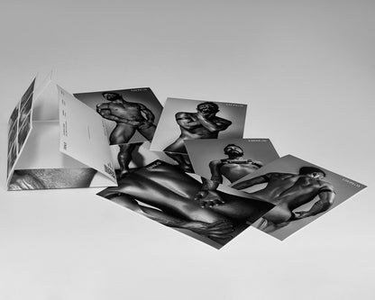 Sobre del sixpack de Lalo (ErosGay) abierto y acostado sobre fondo gris claro, con la pestaña levantada. A un lado, las 6 fotos impresas en blanco y negro están desplegadas y superpuestas en abanico: retratos y cuerpo completo/fragmentos del modelo, todas con estética editorial y alto contraste. Se percibe el set completo: empaque + seis imágenes sueltas