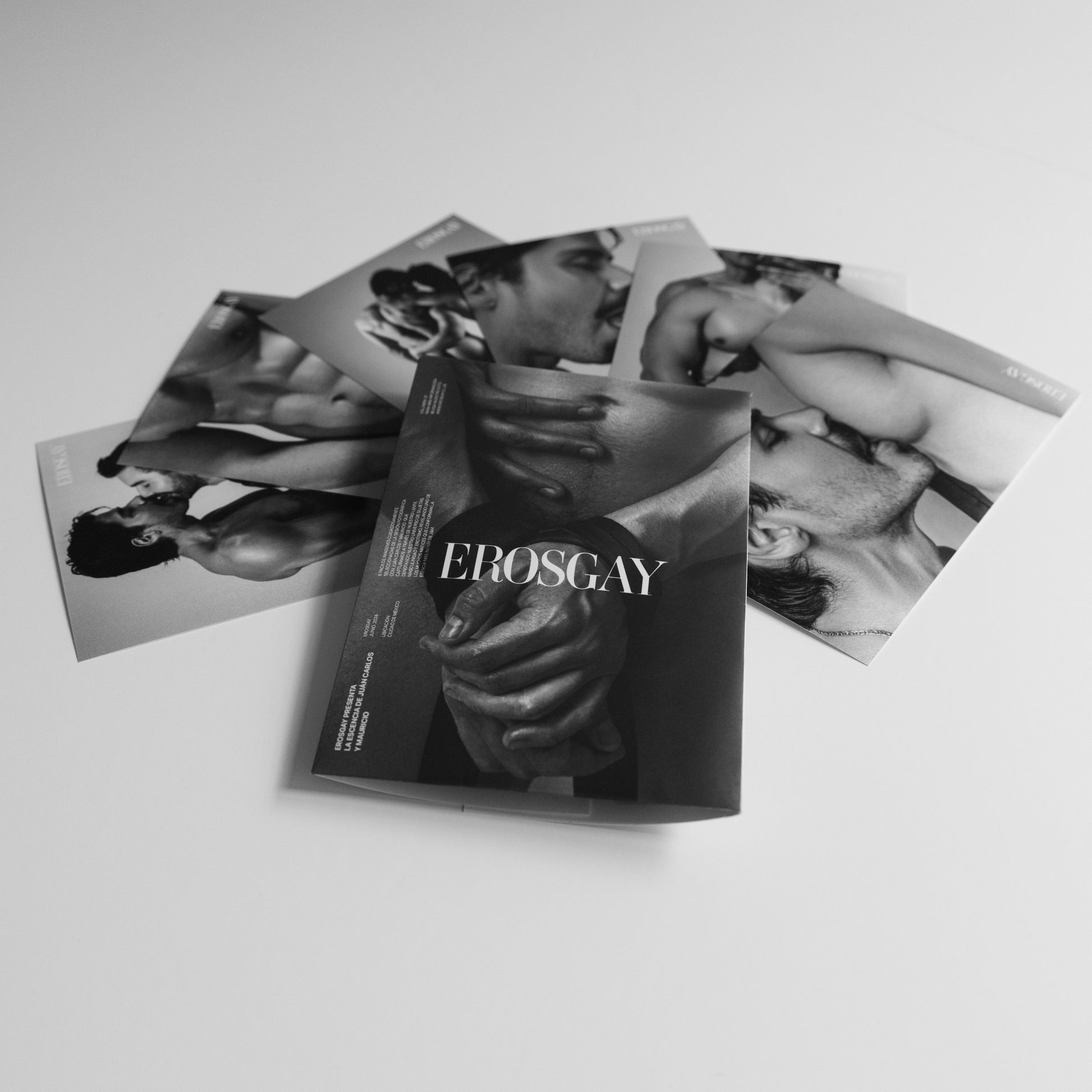 Sobre del Sixpack de ErosGay en blanco y negro con el logo al centro, colocado al frente; detrás, las seis fotos del sixpack están extendidas y parcialmente superpuestas sobre una superficie clara.
