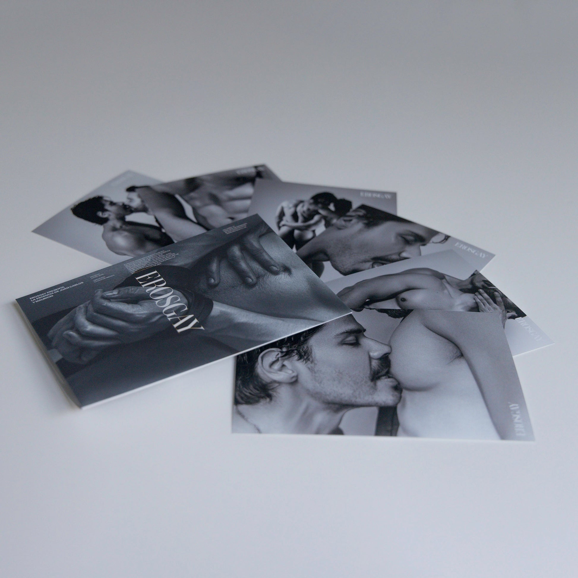 Sobre del Sixpack de ErosGay en blanco y negro con el logo al centro, colocado al frente; detrás, las seis fotos del sixpack están extendidas y parcialmente superpuestas sobre una superficie clara.
