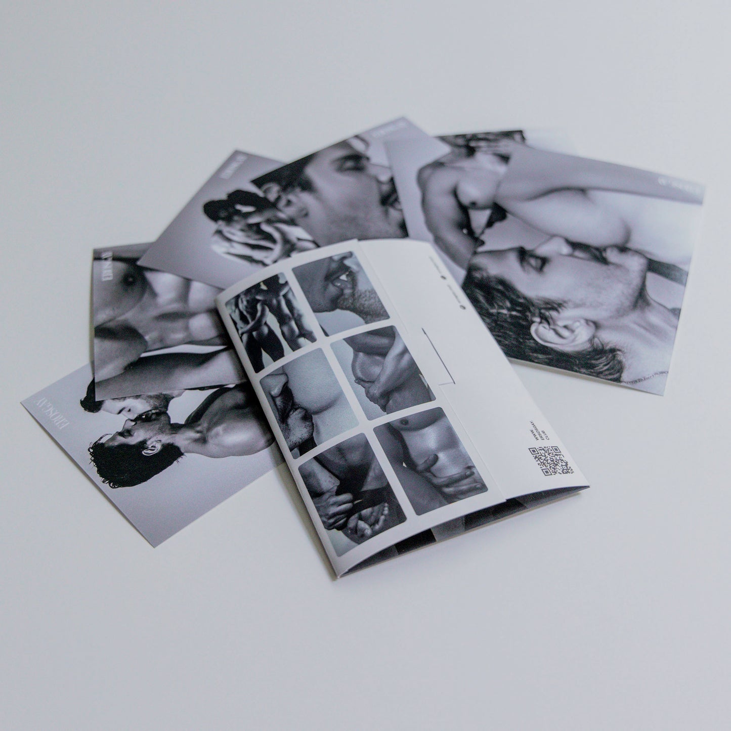 Vista trasera del sobre del Sixpack de ErosGay en blanco y negro, abierto y colocado sobre una superficie clara; a un lado se ven las seis fotos del sixpack extendidas y superpuestas, con detalles del reverso del sobre y un código QR visible.