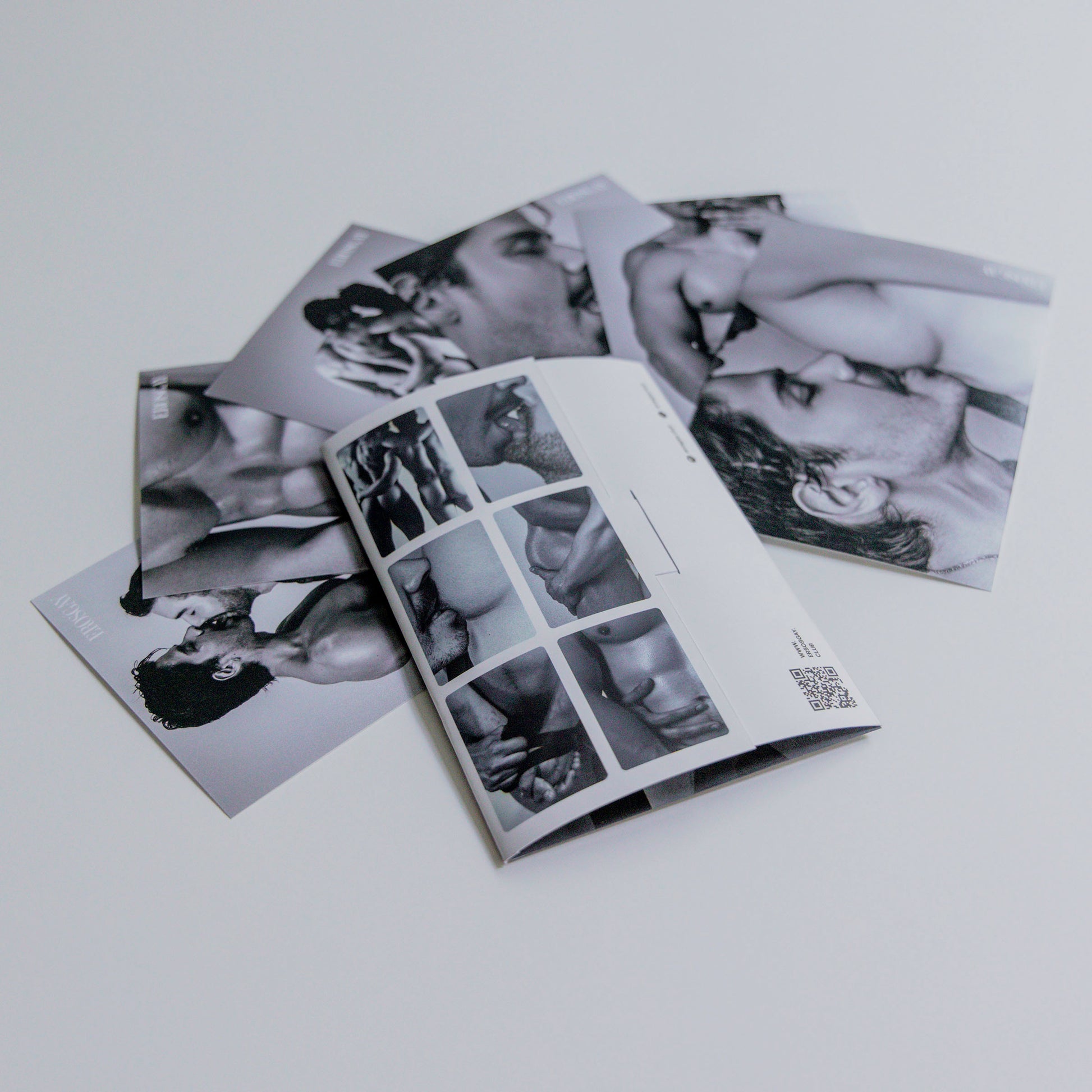 Vista trasera del sobre del Sixpack de ErosGay en blanco y negro, abierto y colocado sobre una superficie clara; a un lado se ven las seis fotos del sixpack extendidas y superpuestas, con detalles del reverso del sobre y un código QR visible.