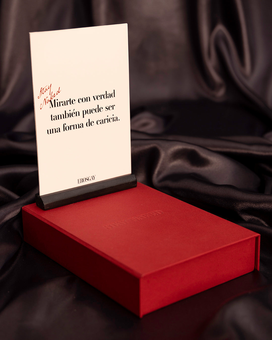 sobre fondo de tela satinada de color negro la caja de color rojo de stay naked red edition con una tarjeta puesta en la base de color negro de la misma coleccion, en la tarjeta se lee la frase stay naked, mirarte con verdad tambien puede ser una forma de caricia.